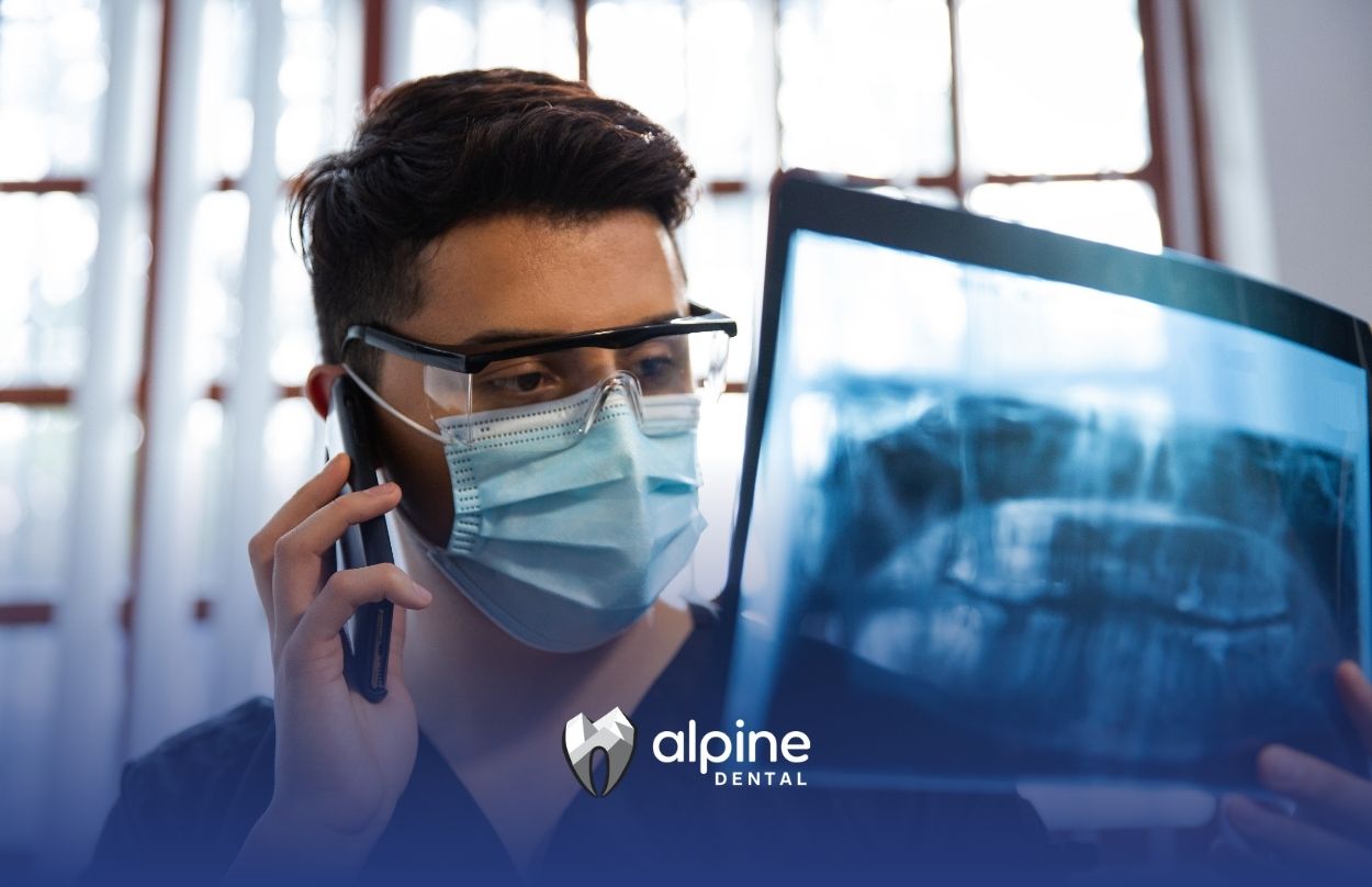 Dental professional wearing protective equipment while examining an X-ray image on a monitor during a patient consultation.
